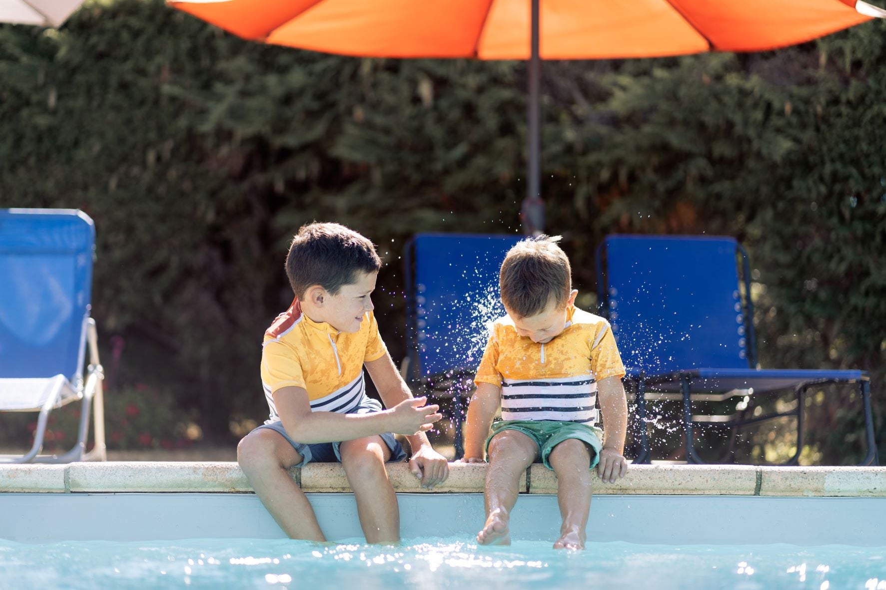 Due ragazzi con magliette a prova di annegamento seduti a bordo piscina che si spruzzano l'un l'altro con l'acqua.